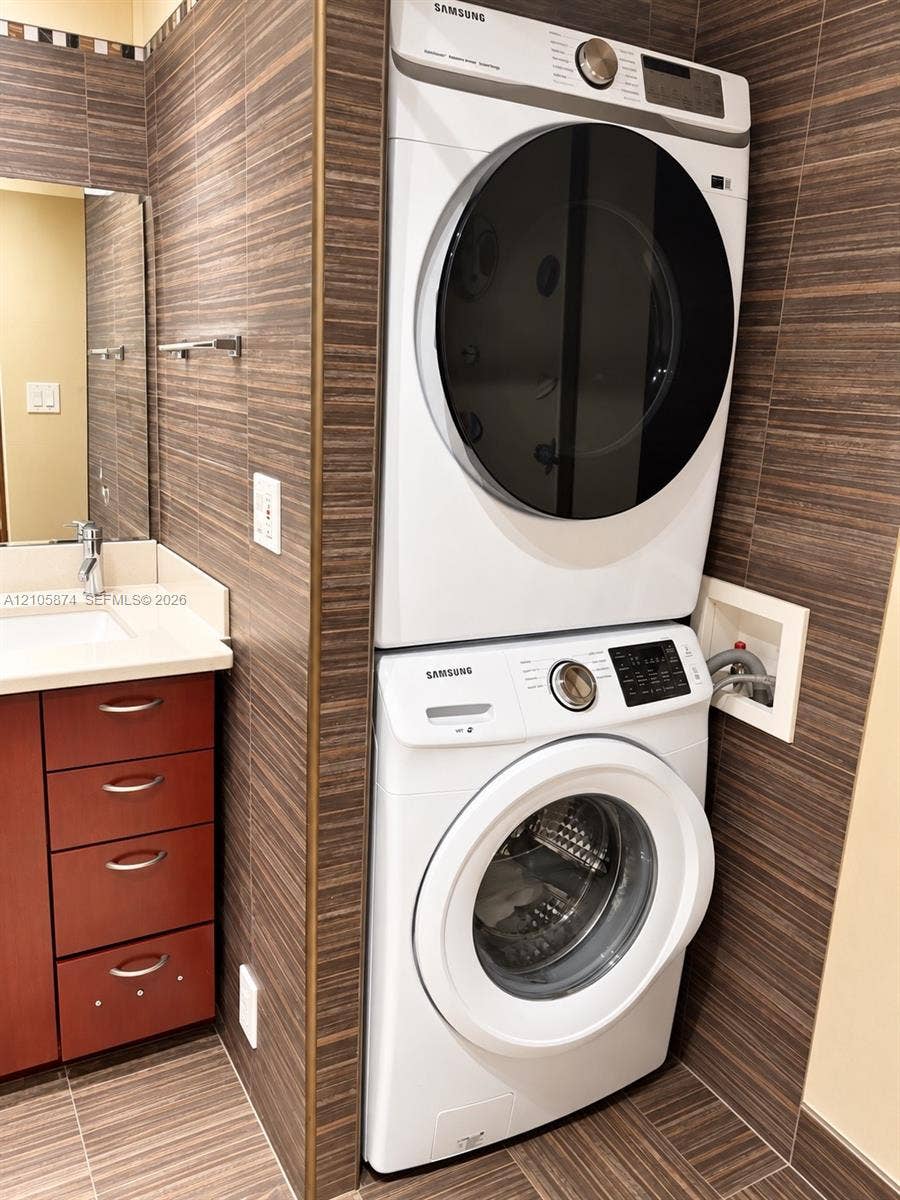 Stacked front-load laundry unit set against floor-to-ceiling textured tile wall, paired with a vanity featuring a solid surface countertop, rectangular undermount sink, and cherry-finish cabinetry