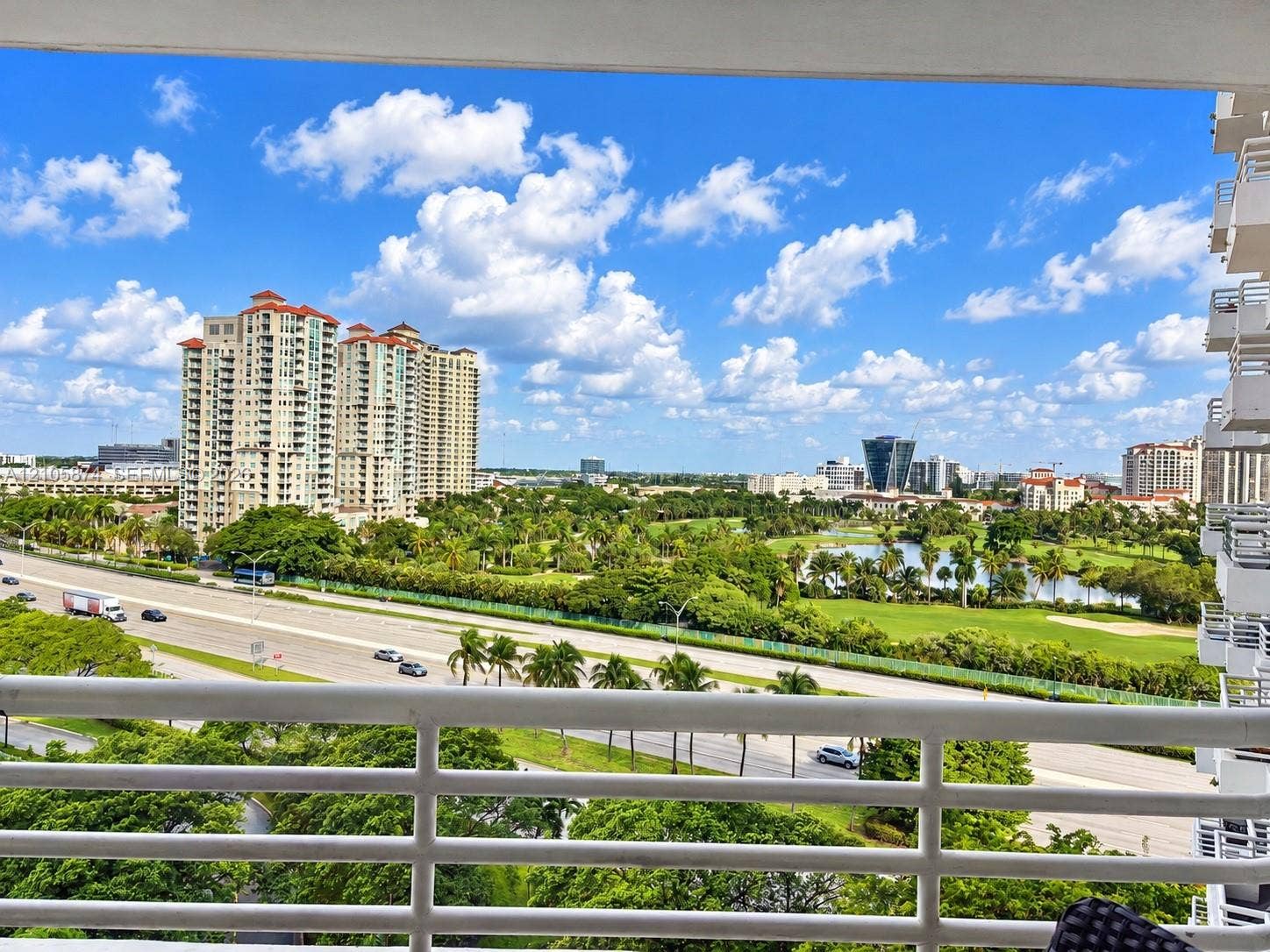 Private balcony with white horizontal railing overlooking expansive greenspace, mature landscaping, and regional cityscape