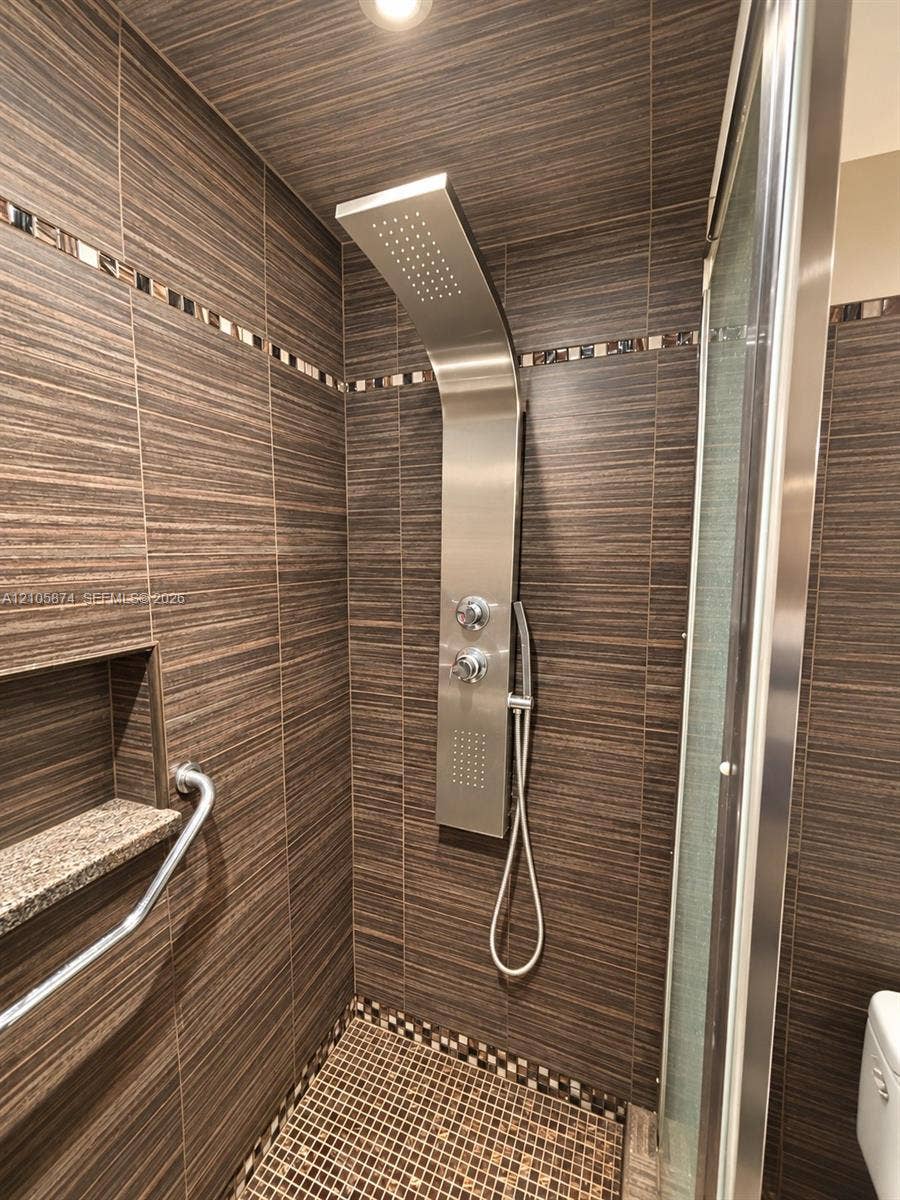 Walk-in shower featuring textured wall tile, a stainless steel rainfall shower panel with a handheld wand, a stone-topped recessed niche, and mosaic tile accents