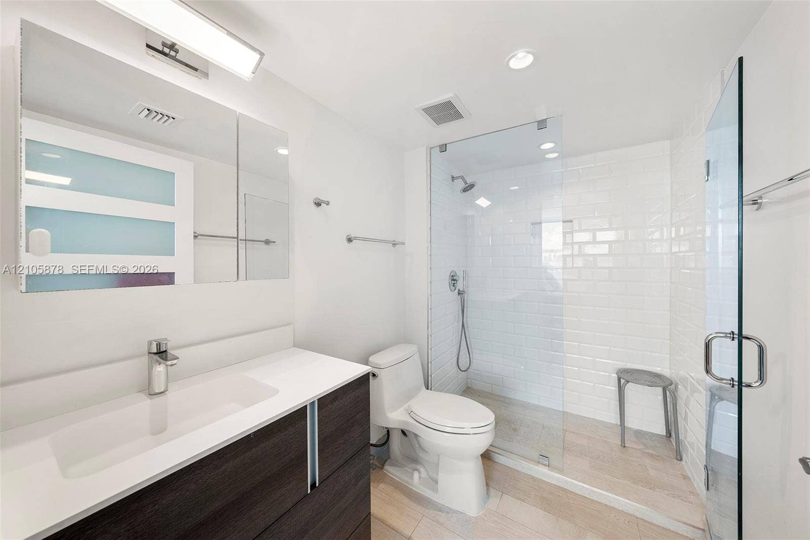 650 West Avenue, #1807, Miami Beach, FL 33139 Dark-finish floating vanity with a white countertop and rectangular integrated sink, walk-in shower with white subway tile surround and glass enclosure, wall-mounted medicine cabinet with integrated lighting, and light-toned tile flooring