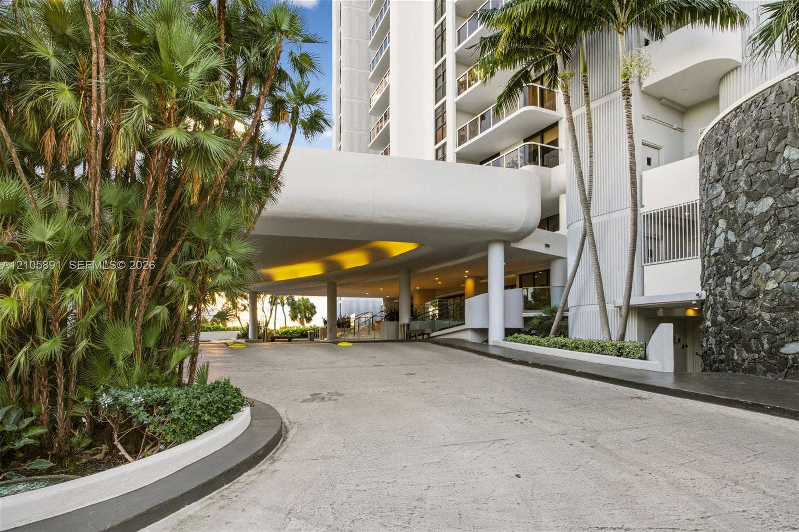 9 Island Avenue, #1215, Miami Beach, FL 33139 Covered porte-cochre entry featuring stone masonry accents, a concrete drive, and mature palm landscaping