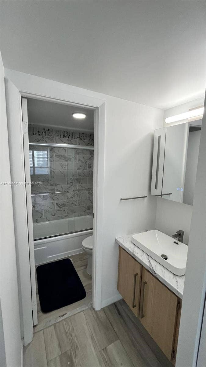 600 Three Islands Boulevard, #913, Hallandale Beach, Hallandale Beach, FL 33009 Vanity featuring a wood-finish cabinet, stone-patterned countertop, and rectangular vessel sink, complemented by wood-finish flooring and a tiled bathtub surround