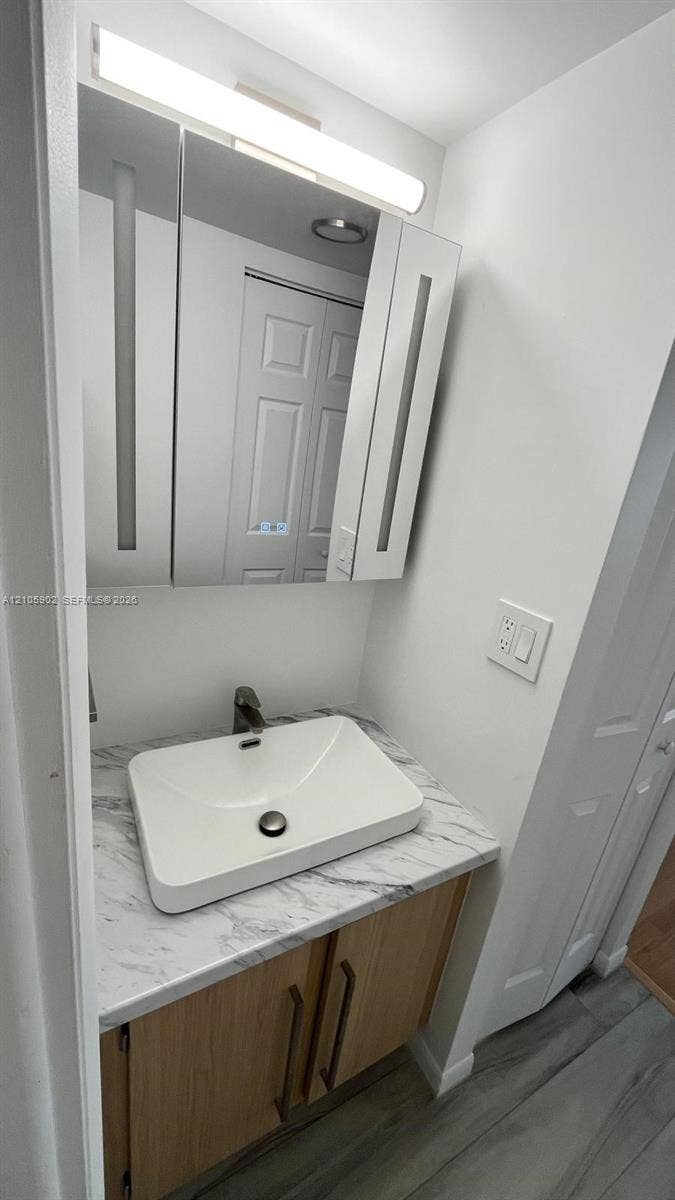 600 Three Islands Boulevard, #913, Hallandale Beach, Hallandale Beach, FL 33009 Vanity featuring a stone-finish countertop, rectangular vessel sink, wood-tone cabinetry with vertical hardware, and an integrated mirrored medicine cabinet with linear lighting