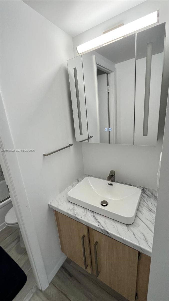 600 Three Islands Boulevard, #913, Hallandale Beach, Hallandale Beach, FL 33009 Vanity featuring a wood-finish cabinet, stone-patterned countertop, vessel sink, integrated medicine cabinet with mirrored doors, and an overhead light fixture