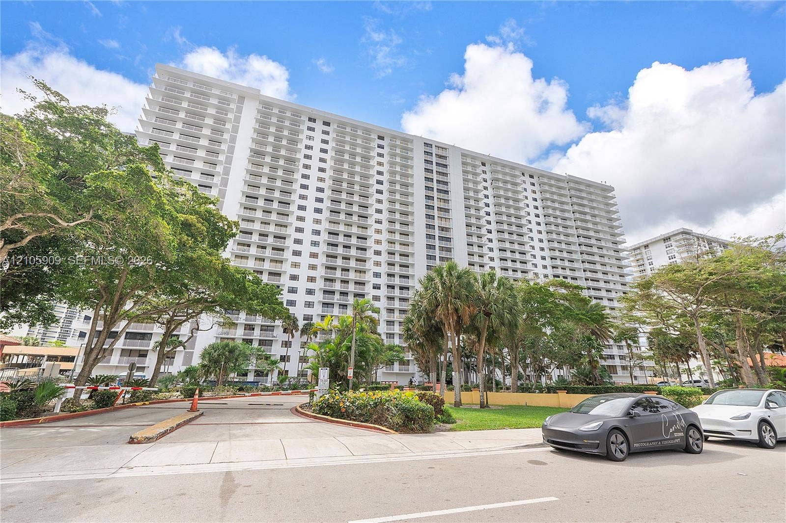 High-rise residential exterior featuring private balconies, concrete construction, paved vehicular entrance, and mature landscaping