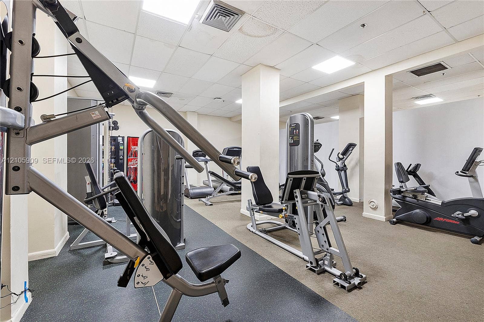 Fitness room featuring a drop ceiling, recessed lighting, structural support columns, and industrial-grade rubber flooring
