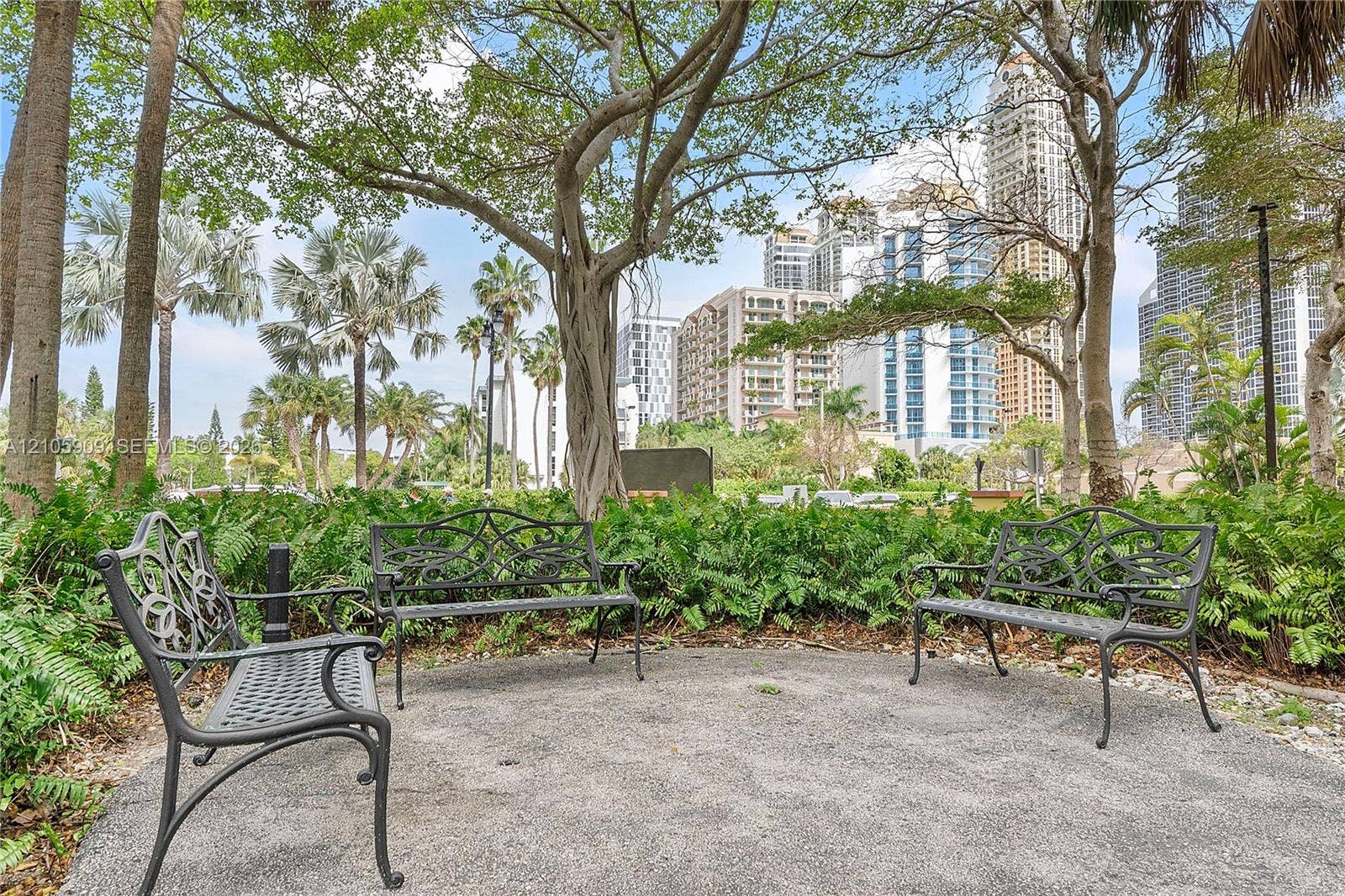 Landscaped grounds featuring crushed stone pathways, mature native trees, tropical foliage, and decorative wrought-iron benches