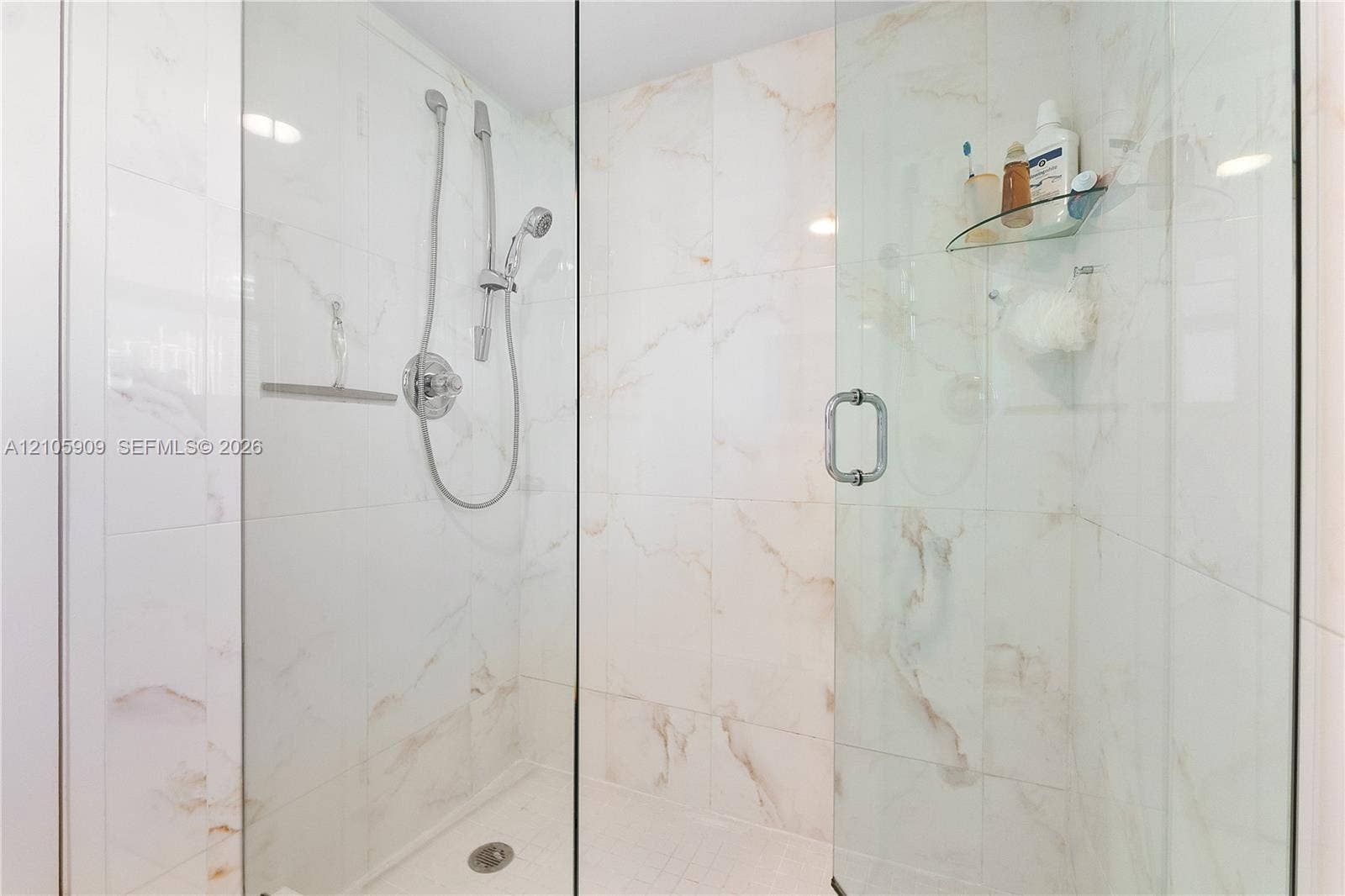 Walk-in shower featuring large-format stone-finish tile surround, frameless glass enclosure, adjustable hand-held showerhead, and integrated glass corner shelving