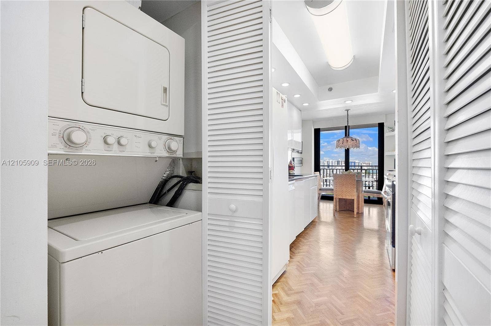 Stacked laundry unit housed within a closet featuring white louvered doors, situated off a kitchen area with herringbone-patterned wood-finish flooring, recessed lighting, and large windows