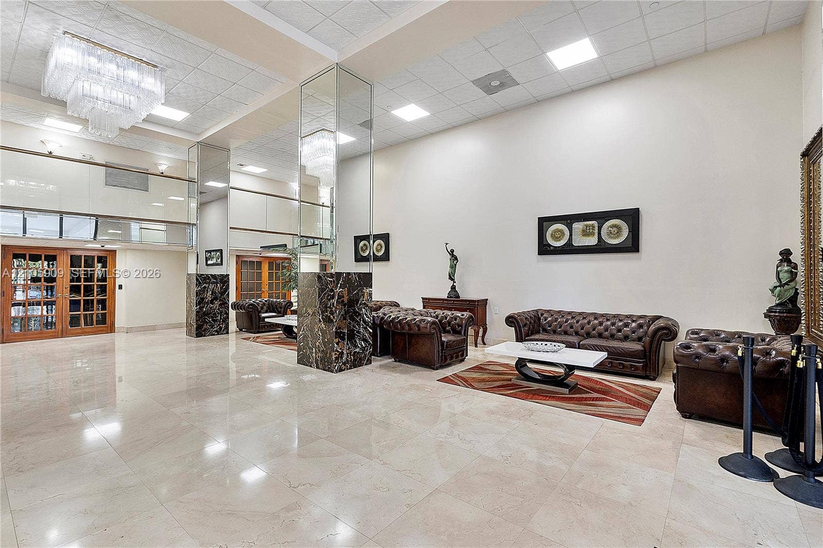 Expansive lobby featuring polished stone flooring, high ceilings with recessed lighting, tiered crystal chandelier, and mirrored support columns with dark stone bases