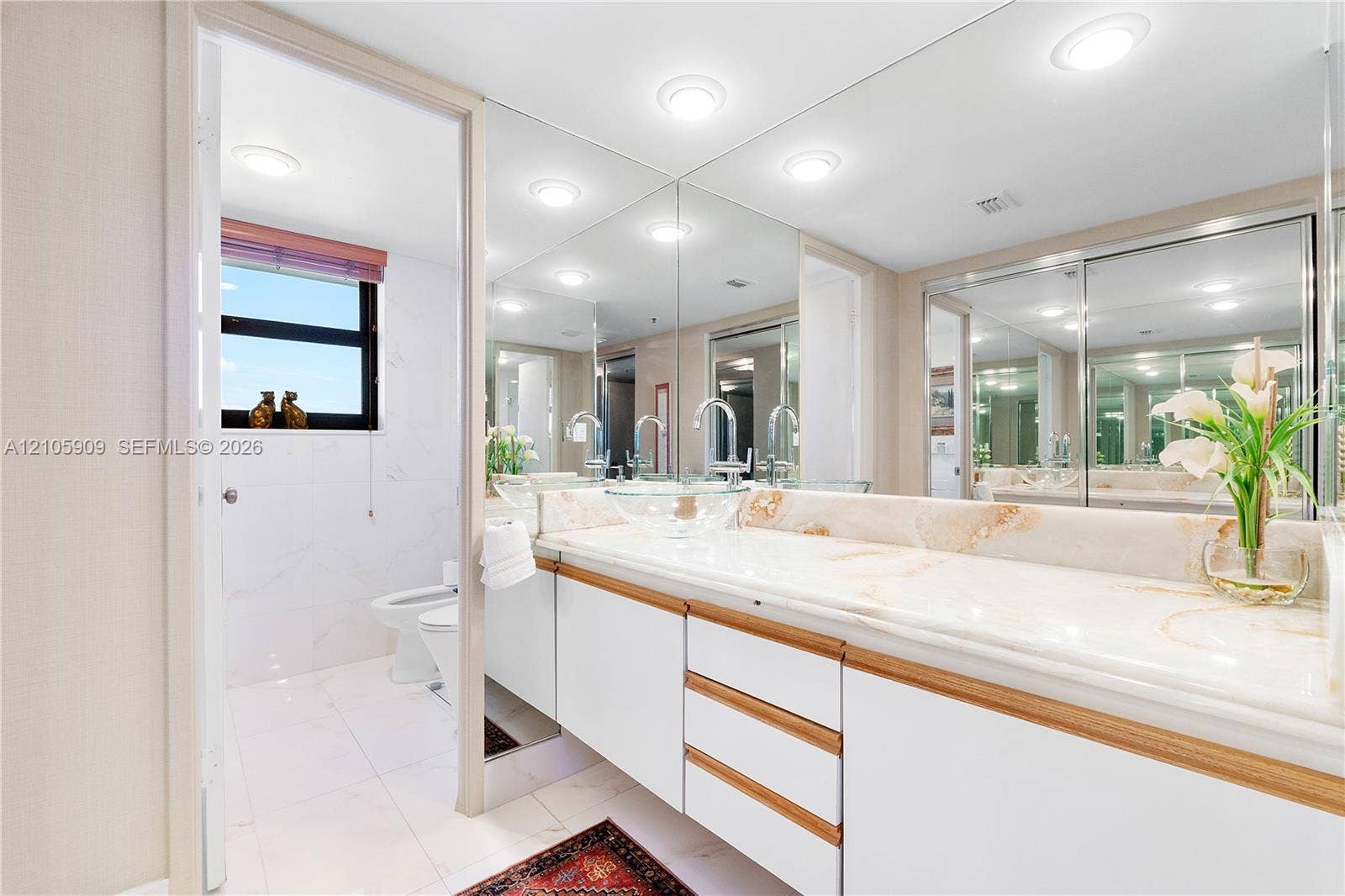 Full-width vanity featuring white cabinetry with wood-tone accents, stone-surface countertop, recessed ceiling lighting, and expansive wall-to-wall mirror