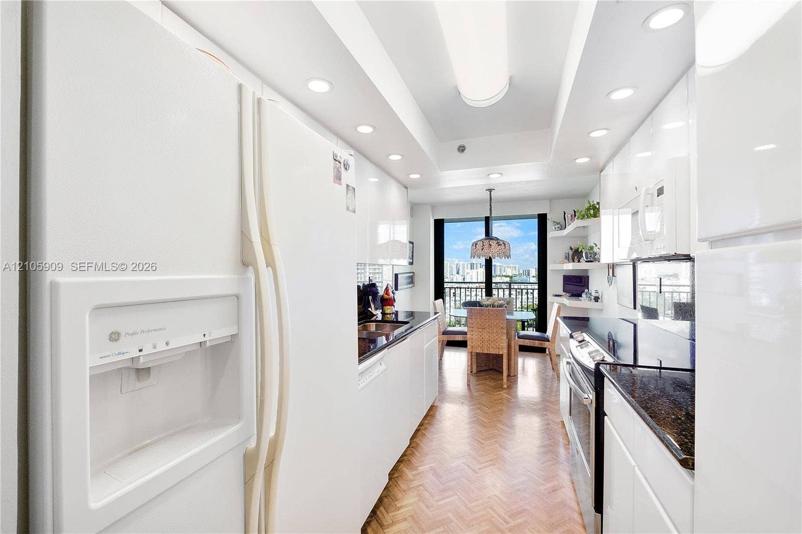 Galley kitchen featuring high-gloss white cabinetry, dark stone countertops, herringbone wood-finish flooring, recessed ceiling lighting, and built-in stainless steel appliances