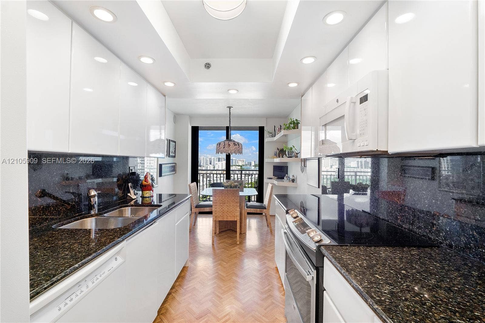 Galley kitchen featuring high-gloss white cabinetry, dark stone countertops, herringbone patterned wood flooring, recessed lighting, and built-in white appliances