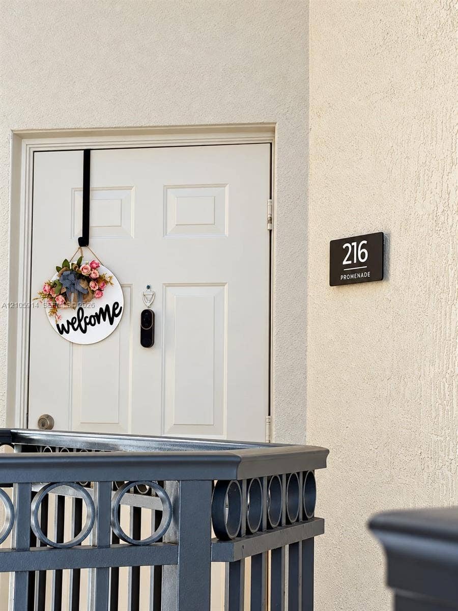 11102 NW 83rd Street, #216, Doral, FL 33178 White paneled entry door with integrated peephole and smart doorbell camera, complemented by a matte finish metal railing featuring circular decorative detailing and a mounted address plaque