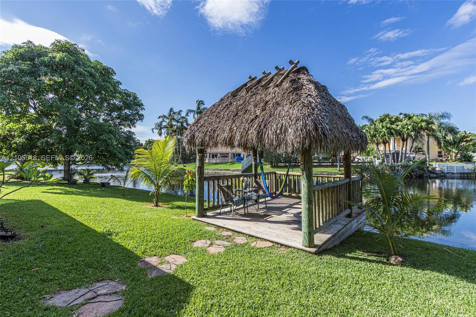 Waterfront tiki-style gazebo featuring a thatched roof, timber structural posts, and wooden decking set upon a grass lawn with stone pavers
