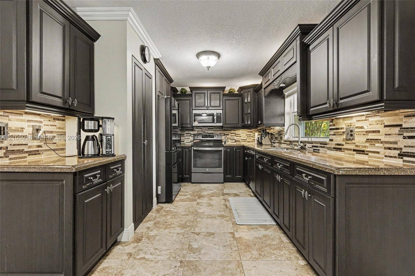Kitchen featuring dark-finish cabinetry, stone-texture backsplash, neutral stone-finish flooring, stainless steel appliances, and crown molding