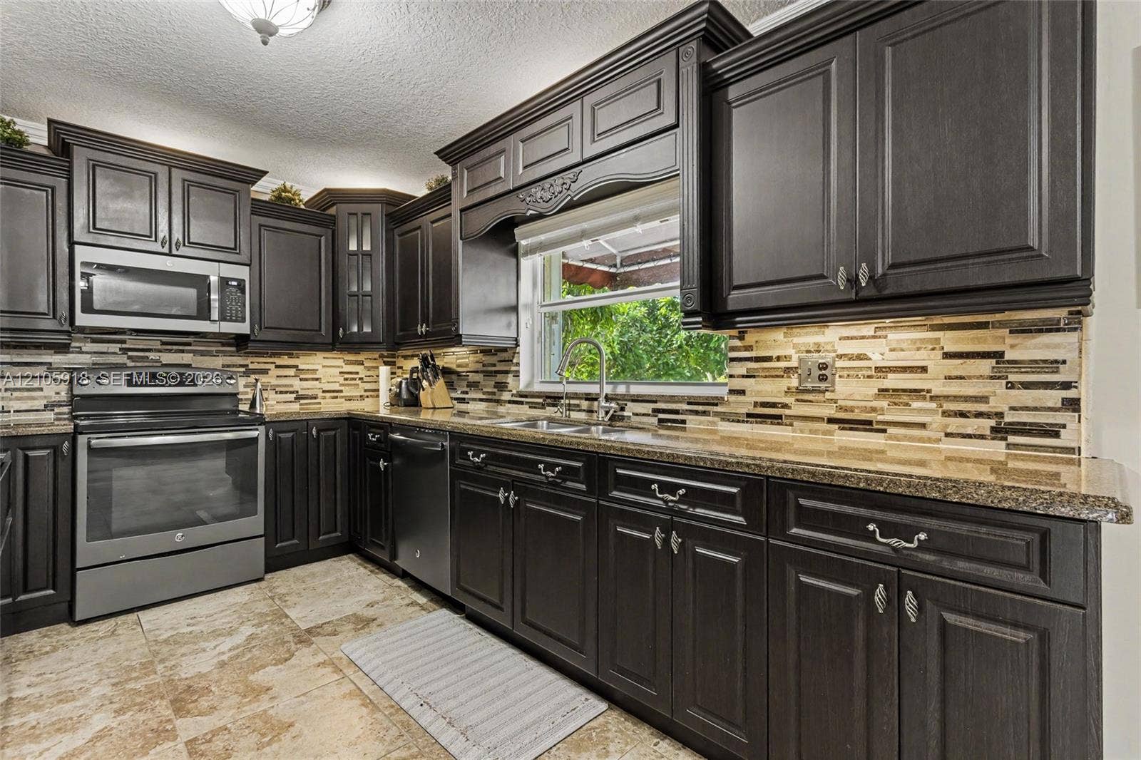 Kitchen featuring dark cabinetry with crown molding, stone-pattern tile flooring, multi-tonal linear backsplash, granite countertops, and stainless steel appliances
