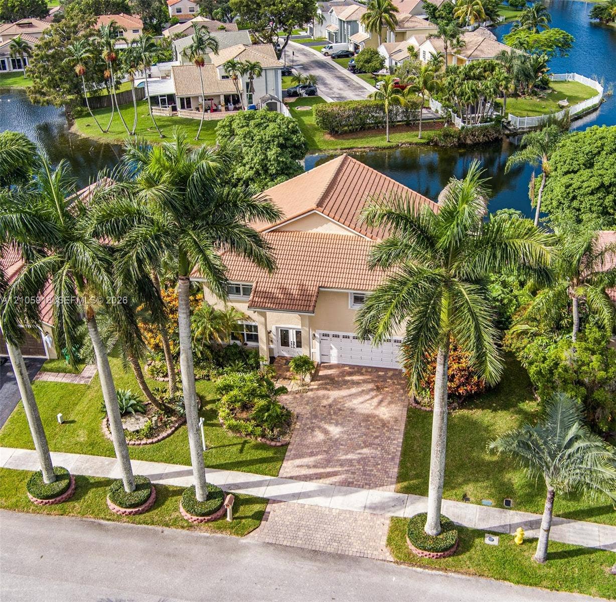 Two-story residence featuring a tile roof, brick-paver driveway, multi-car garage, mature palm landscaping, and waterfront positioning