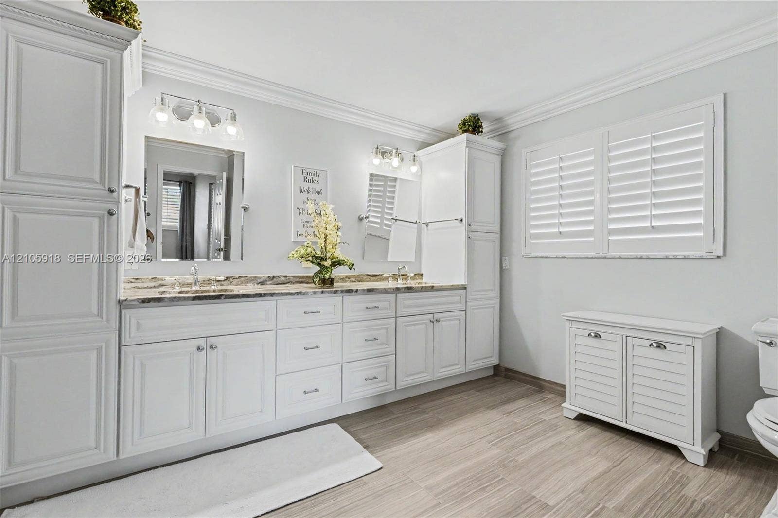 Double vanity featuring white cabinetry, stone-finish countertops, dual mirrors with integrated lighting, and plantation-style window shutters
