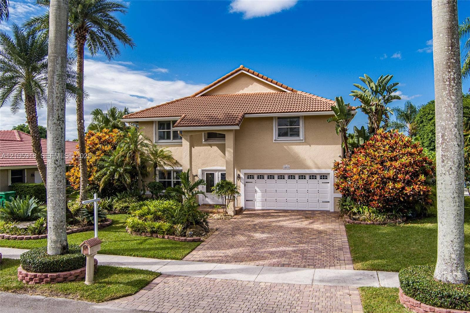 Two-story residence featuring a Mediterranean-style clay tile roof, stucco exterior, paver driveway, attached two-car garage, and established tropical landscaping