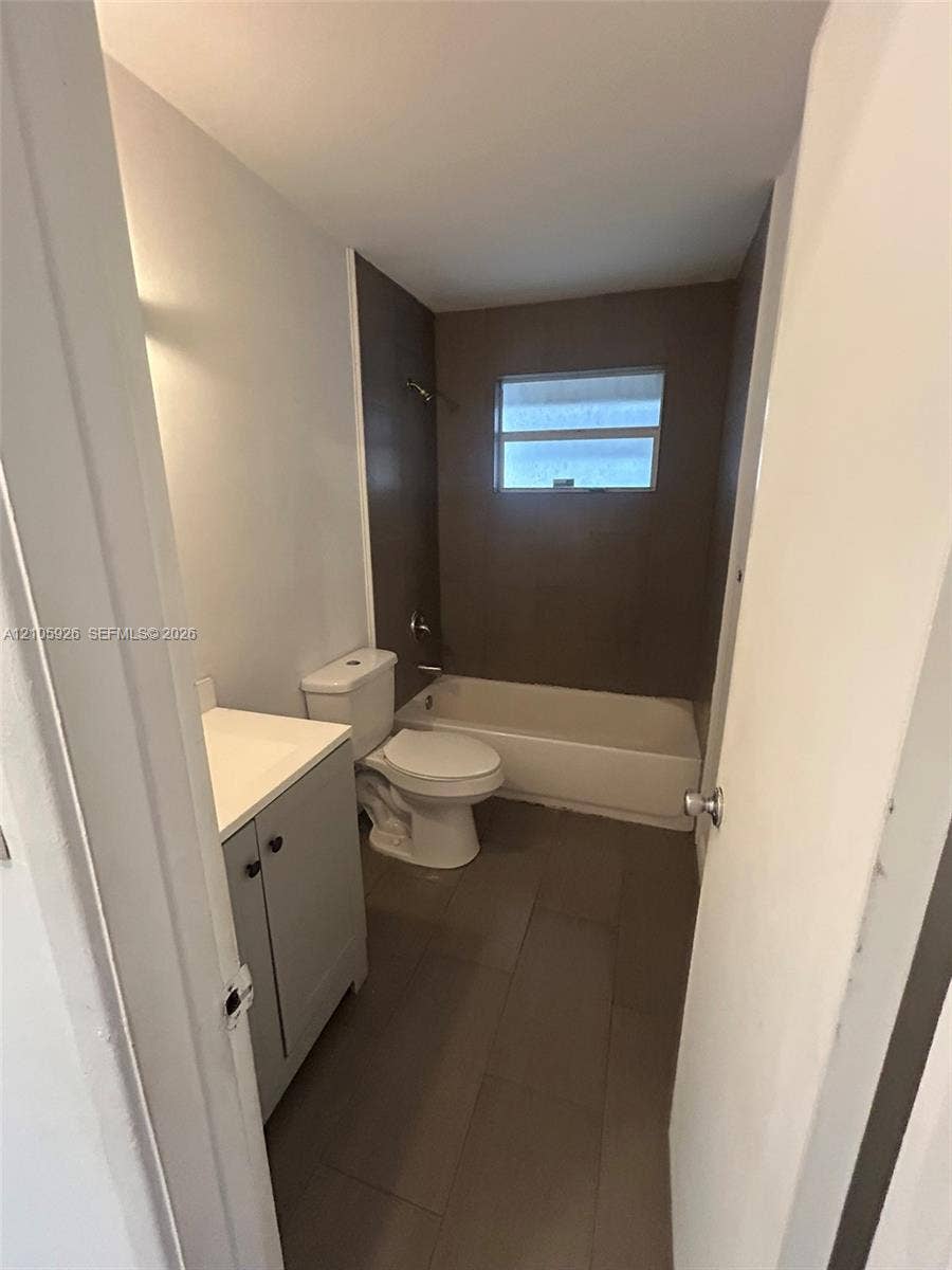 1601 W Avenue H W, Riviera Beach, Riviera Beach, FL 33404 Bathroom featuring large-format tile flooring, a vanity with a white countertop, a porcelain toilet, and a combination tub and shower enclosure with dark-toned tile surround