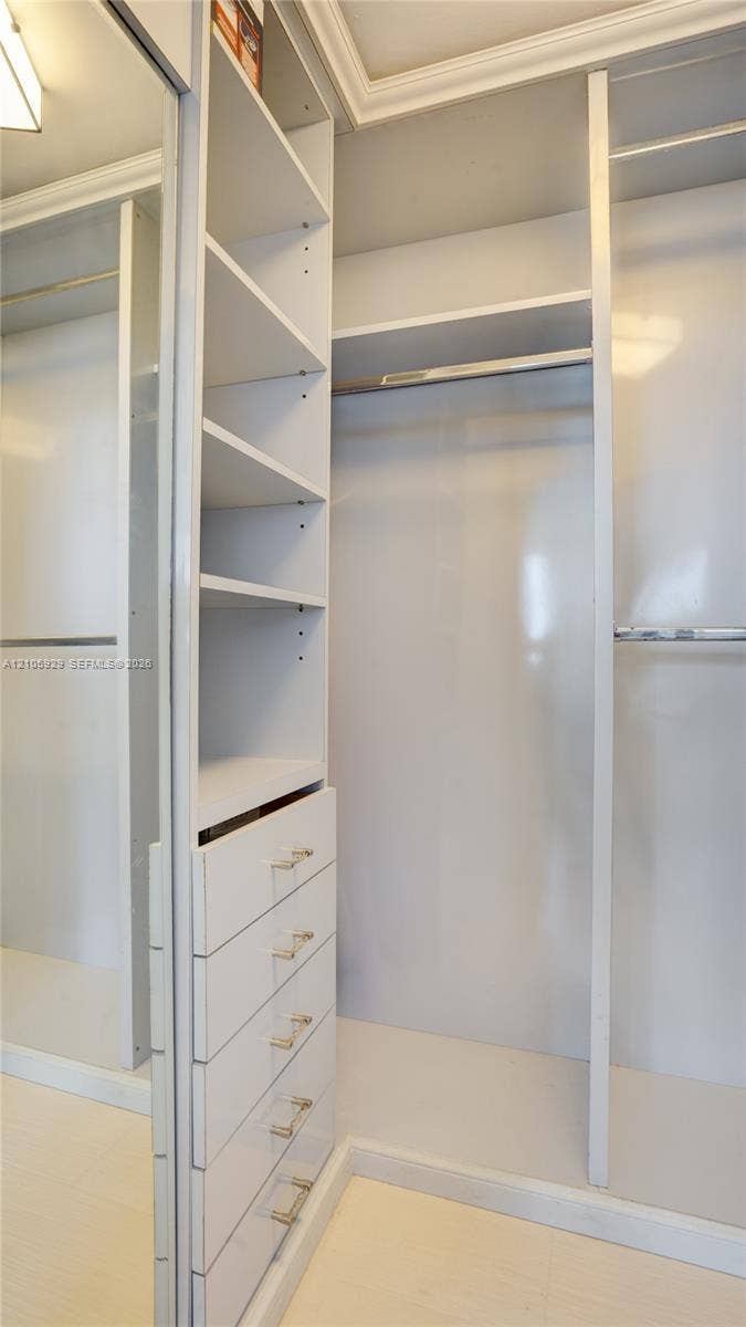 2655 Collins Avenue, #2301, Miami Beach, FL 33140 Custom walk-in closet featuring white built-in shelving, a five-drawer dresser unit with metallic pulls, dual hanging rods, and a full-length mirrored door