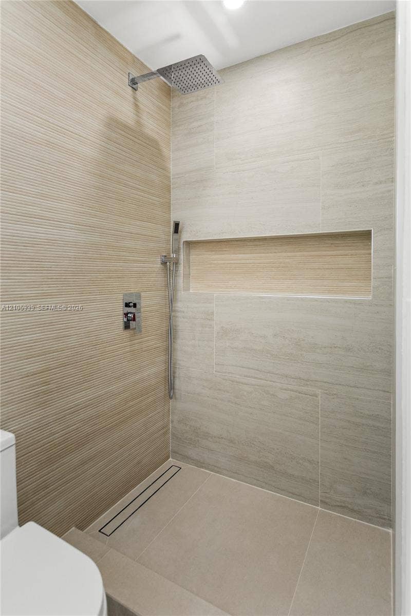 2850 NW 205th Street, Miami Gardens, FL 33056 Walk-in shower featuring textured wall tile, large-format smooth wall tile, integrated recessed shelving, square rainfall showerhead with handheld sprayer, and a linear floor drain