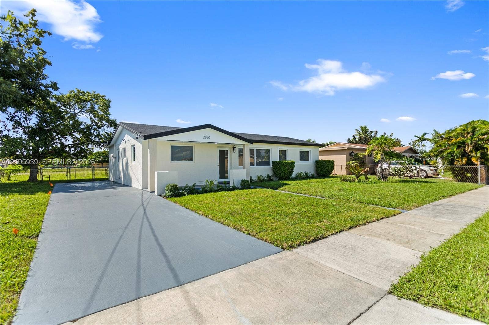 2850 NW 205th Street, Miami Gardens, FL 33056 Single-story exterior featuring light-colored stucco siding, a low-pitched roof, an expansive paved driveway, and manicured front lawn