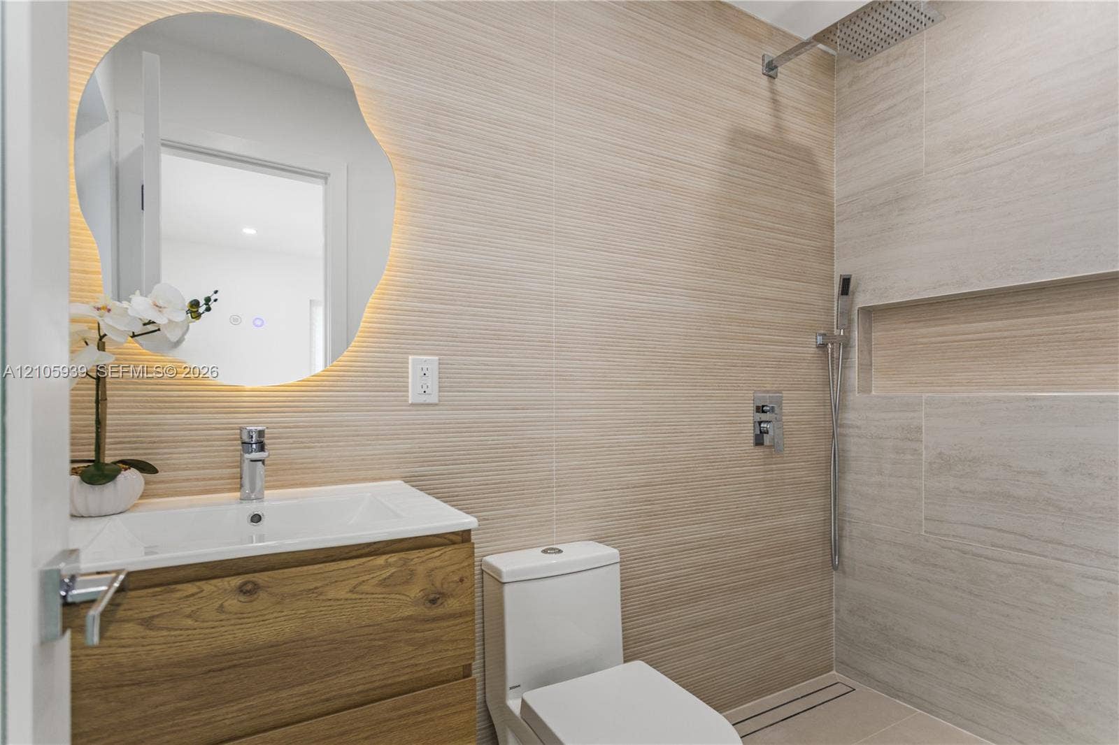 2850 NW 205th Street, Miami Gardens, FL 33056 Wall-mounted wood-finish floating vanity, backlit organic-shaped mirror, horizontal textured wall tile, and walk-in shower featuring a built-in recessed niche and rainfall showerhead