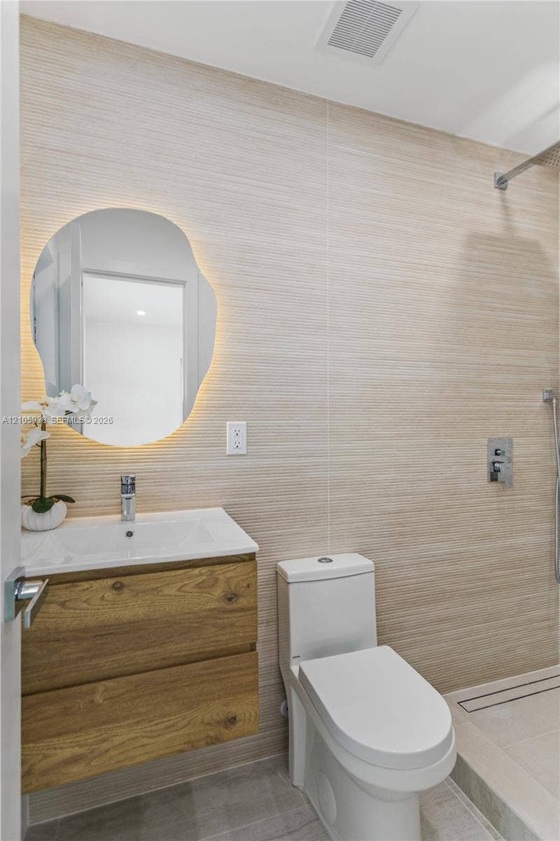 2850 NW 205th Street, Miami Gardens, FL 33056 Floating wood-finish vanity with an integrated sink, backlit organic-shaped mirror, wall-mounted shower fixture, and textured stone-finish wall tile
