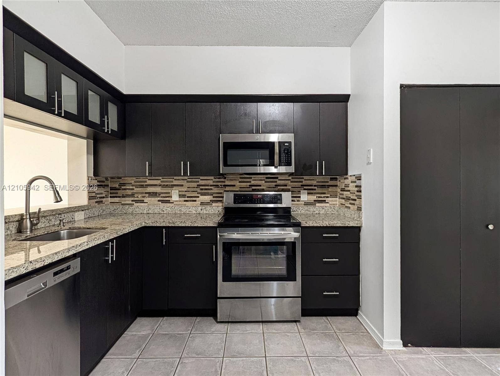 271 Duval Court, Weston, Weston, FL 33326 Dark cabinetry with glass-front upper accents, stone-composite countertops, stainless steel appliances, multi-tonal backsplash, and ceramic tile flooring
