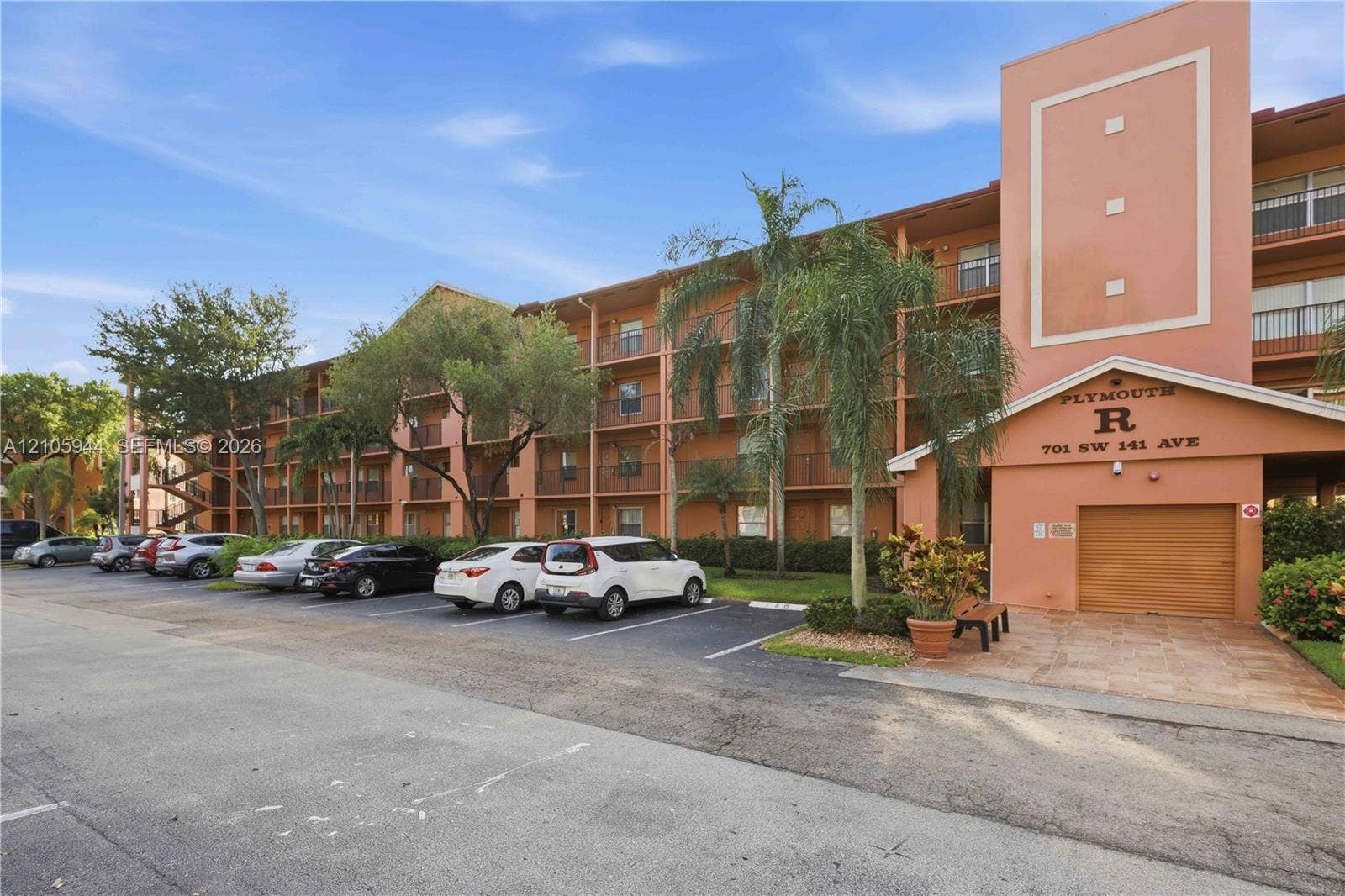 Multi-story building exterior featuring a stucco facade, covered ground-level parking, private balconies with metal railings, and mature palm tree landscaping