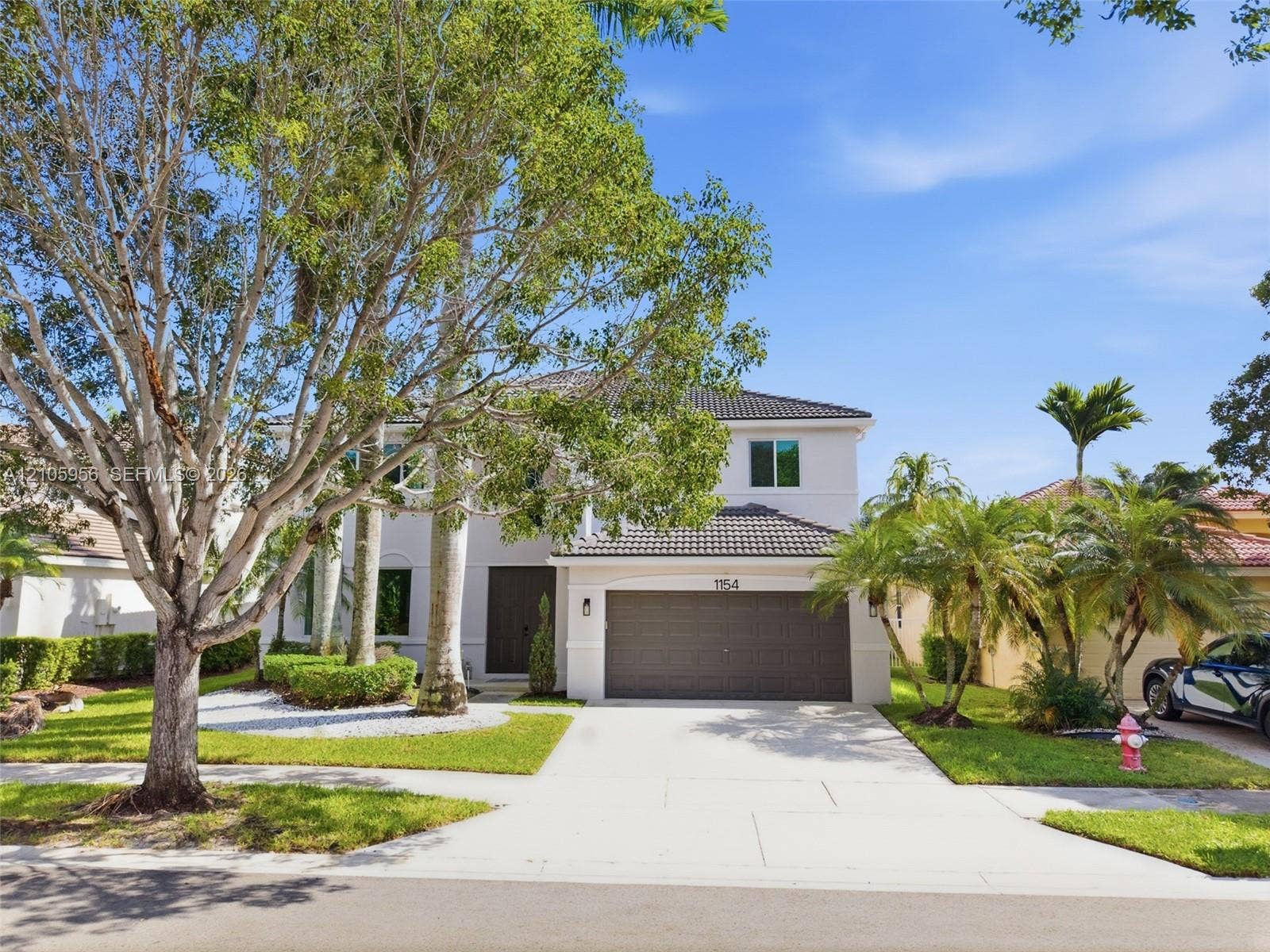 1154 Croton Court, Weston, Weston, FL 33327 Two-story exterior featuring a light-colored stucco facade, tile roofing, an attached two-car garage, and established landscaping with mature trees and palms