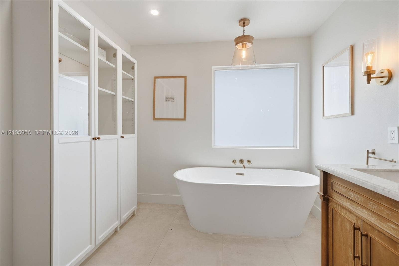 1154 Croton Court, Weston, Weston, FL 33327 Freestanding soaking tub with wall-mounted fixtures, built-in floor-to-ceiling cabinetry with glass-panel doors, neutral tile flooring, wood-finish vanity with stone countertop, and pendant lighting