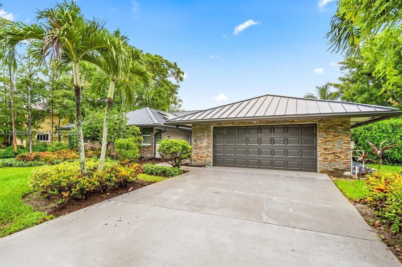 5966 NW 77th Terrace, Parkland, Parkland, FL 33067 Single-story residence featuring a standing seam metal roof, stone veneer exterior, two-car garage, and concrete driveway, accented by established tropical landscaping