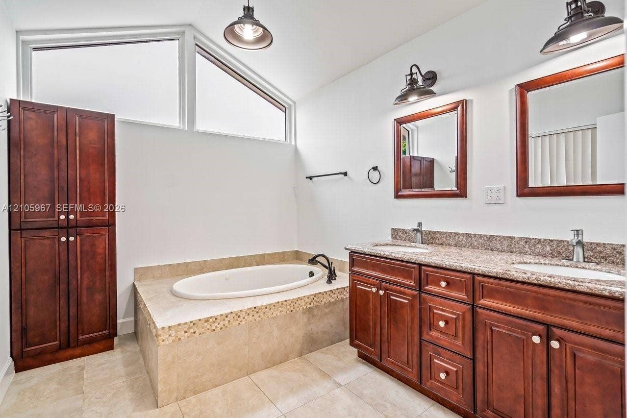 5966 NW 77th Terrace, Parkland, Parkland, FL 33067 Dual-vanity cabinetry with stone-surface countertops, soaking tub with tile surround, vaulted ceiling with transom windows, and wall-mounted metal light fixtures