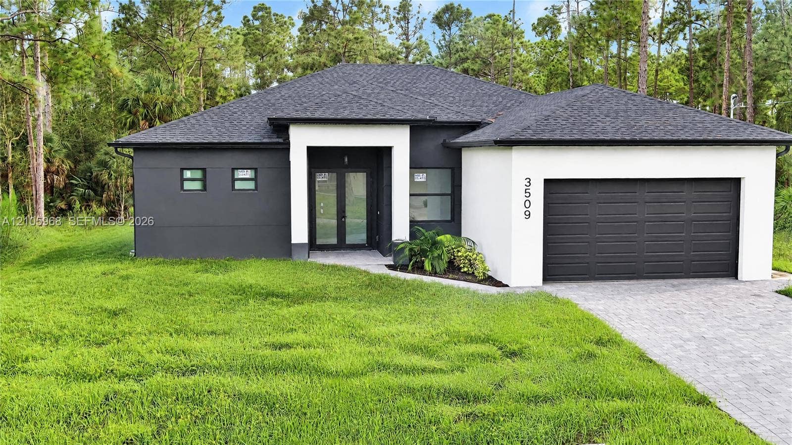 3509 63rd Street W, Lehigh Acres, FL 33971 Modern single-story exterior featuring high-contrast stucco facade, stone paver driveway, dual-glass entry doors, and a multi-car garage