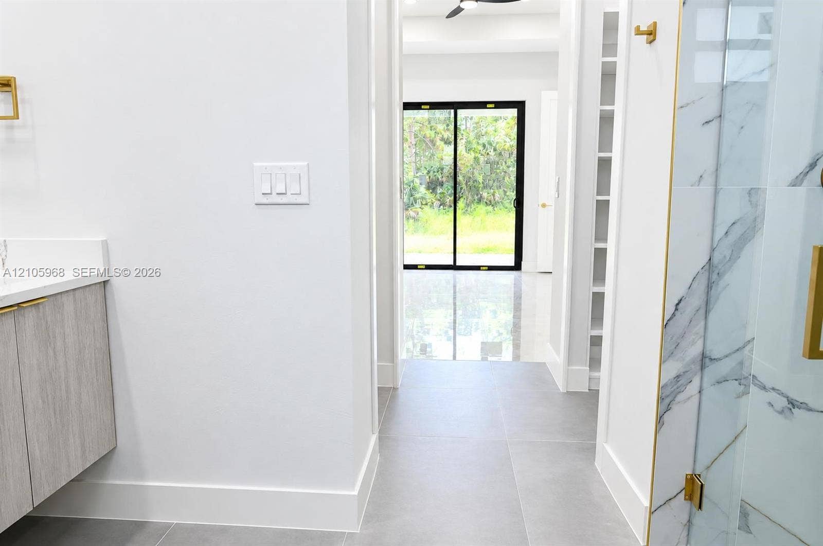 3509 63rd Street W, Lehigh Acres, FL 33971 Large-format gray floor tiles, vanity with textured wood-finish cabinetry, walk-in shower with stone-patterned tile surround, brass hardware, and floor-to-ceiling built-in shelving