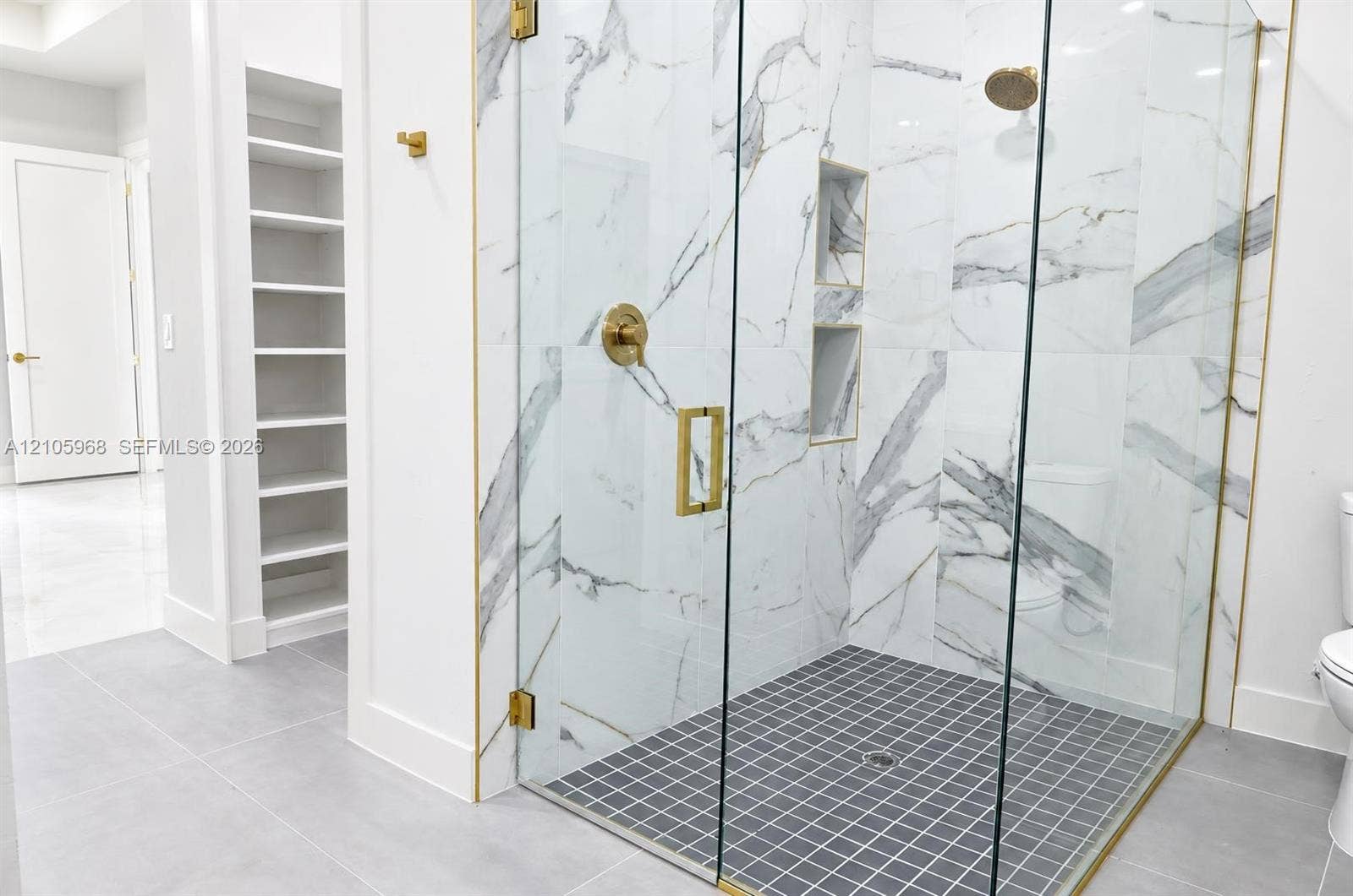 3509 63rd Street W, Lehigh Acres, FL 33971 Walk-in shower featuring gold-toned hardware, floor-to-ceiling tiled walls with marbled pattern, recessed wall shelving, and a glass enclosure, complemented by built-in wall cabinetry
