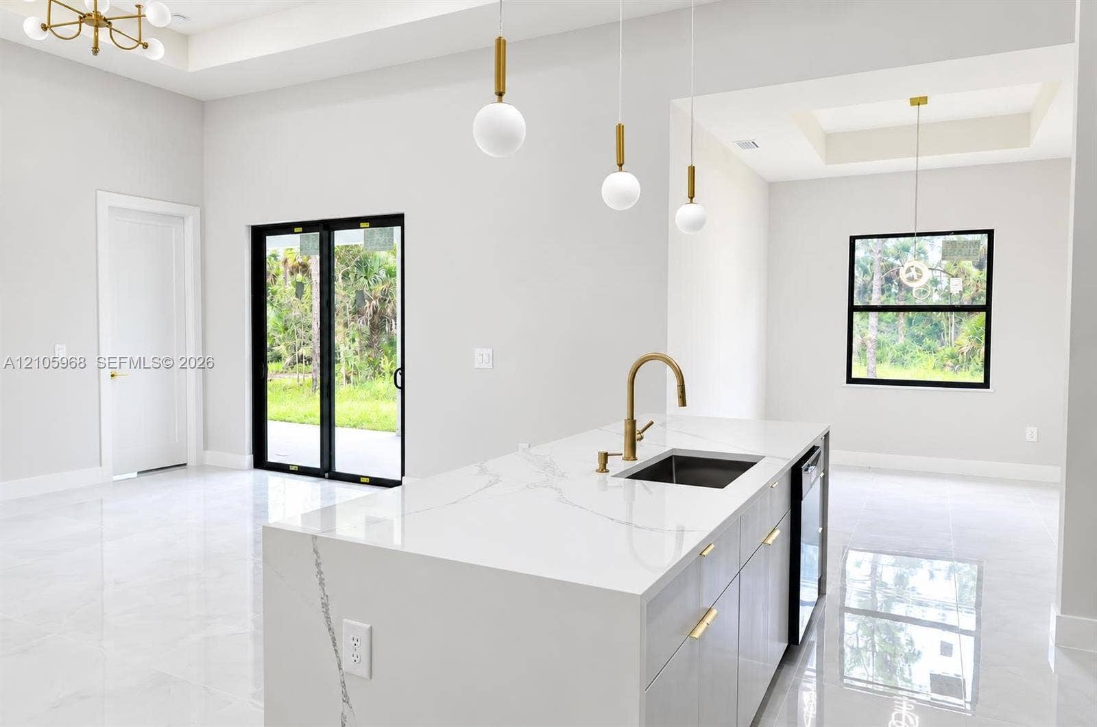 3509 63rd Street W, Lehigh Acres, FL 33971 Kitchen island featuring a waterfall-edge countertop, undermount sink with a brass faucet, contemporary pendant lighting, light-tone cabinetry with gold-finish hardware, and large-format tile flooring