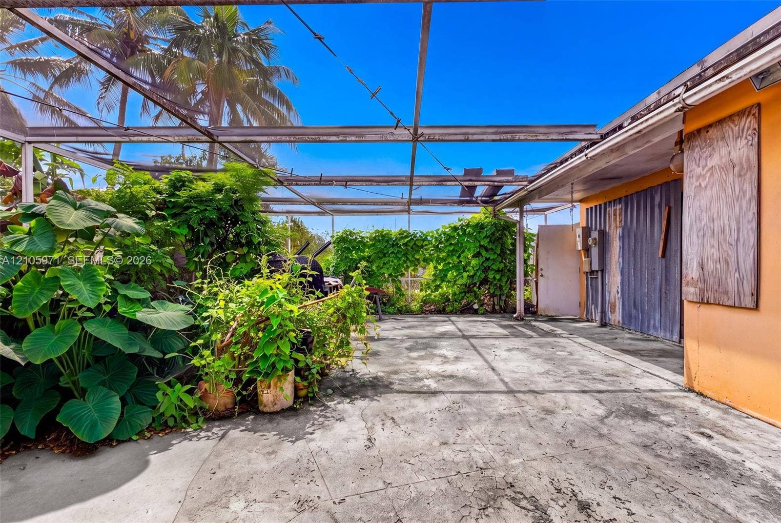 6604 Coconut Drive, Miramar, FL 33023 Screen-enclosed concrete patio featuring established tropical landscaping, metal siding exterior wall accents, and an expansive overhead aluminum frame
