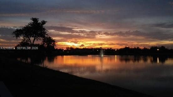 6220 Oceanaire Way, Lake Worth, FL 33467 Waterfront property featuring a lake, active fountain, and mature tree silhouettes set against a sunset backdrop