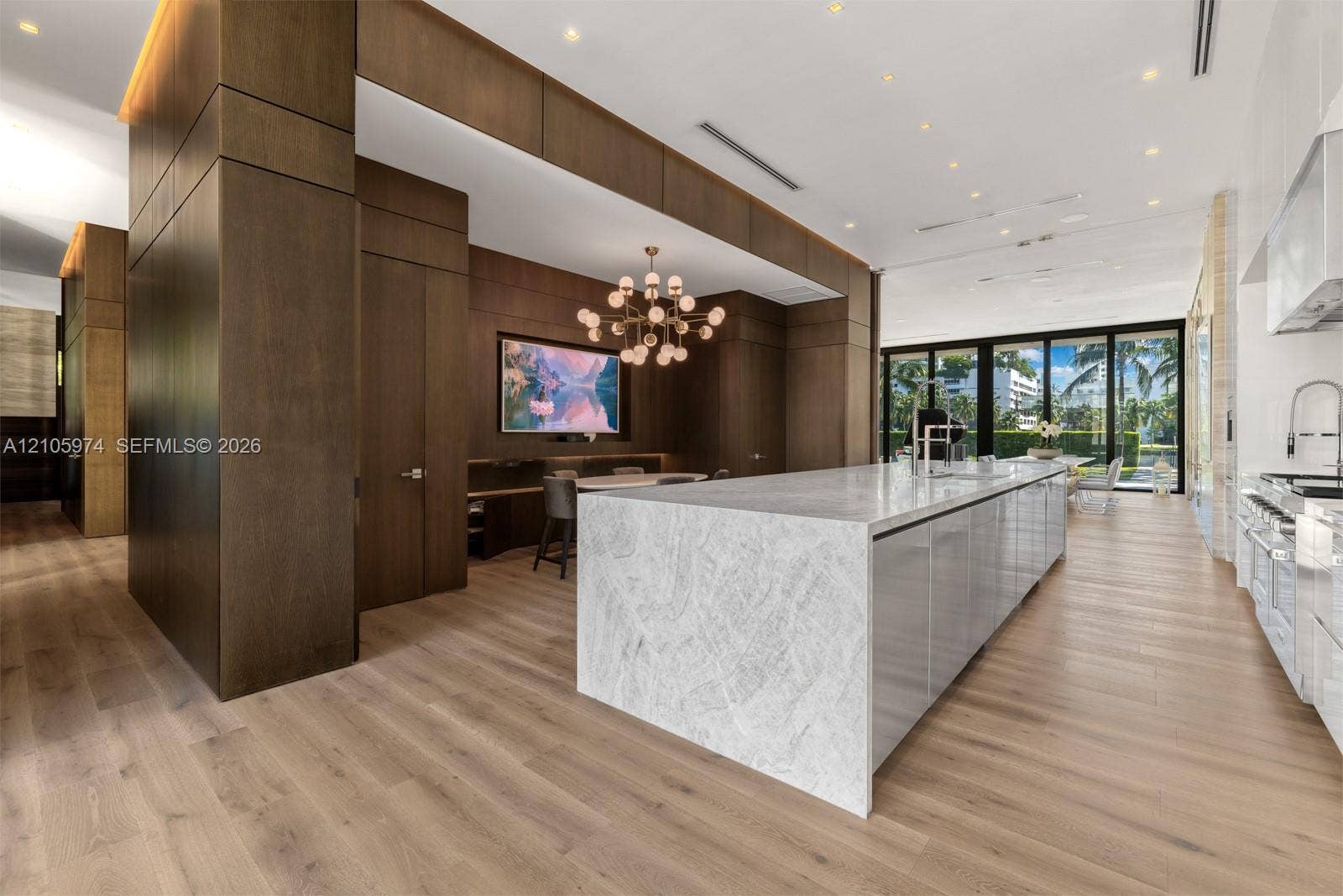 4395 Pine Tree Drive, Miami Beach, FL 33140 Large kitchen featuring a waterfall-edge stone island, wood-panel wall treatments, wood-finish flooring, a multi-globe chandelier, and expansive sliding glass doors