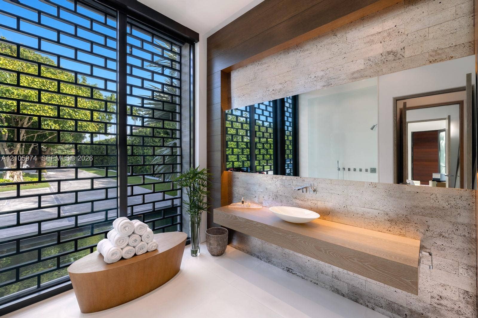 4395 Pine Tree Drive, Miami Beach, FL 33140 Floating wood-finish vanity with a white vessel sink, wall-mounted faucet, oversized frameless mirror, textured stone-tiled wall, and full-height exterior window with integrated metal grid screen