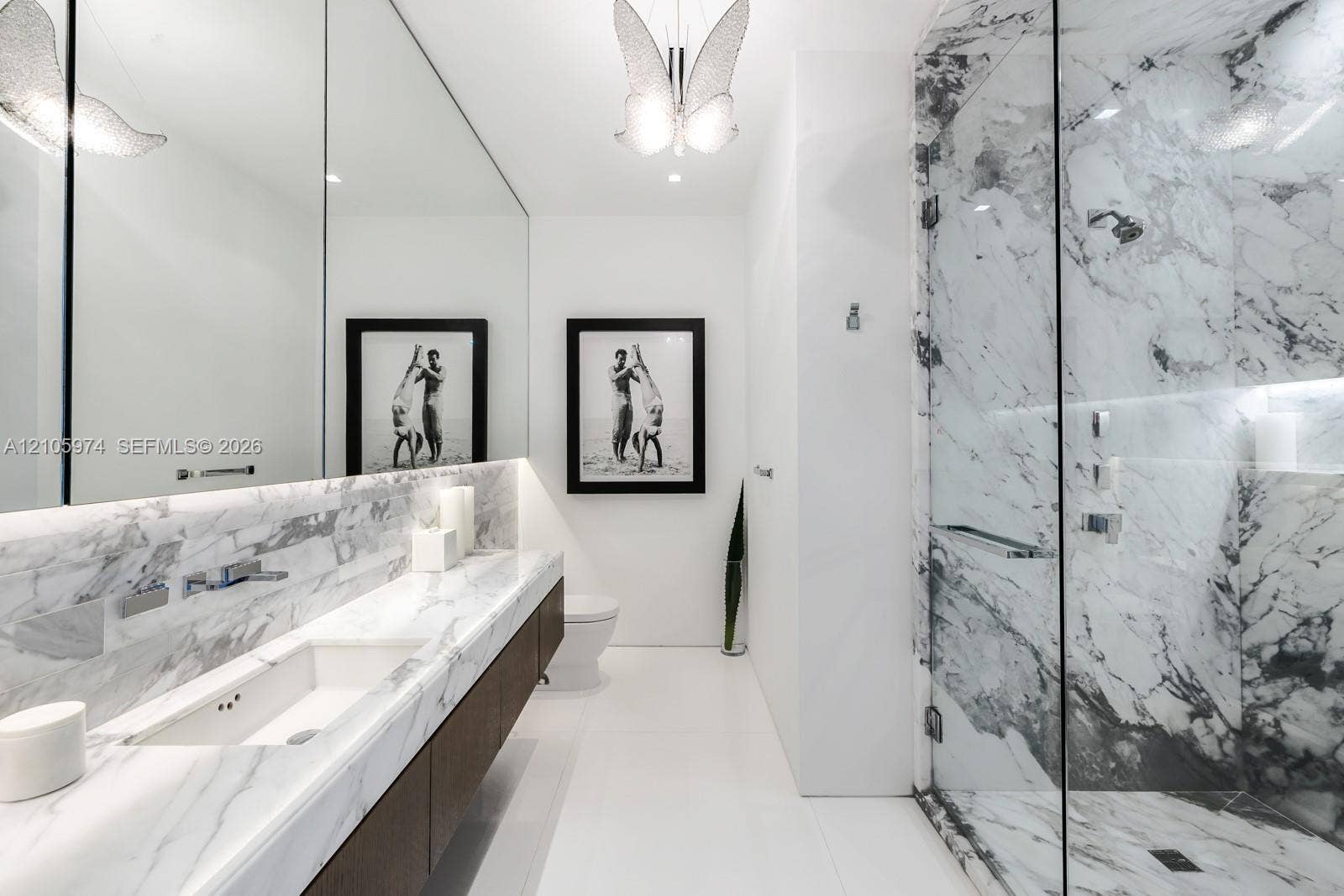 4395 Pine Tree Drive, Miami Beach, FL 33140 Stone-surfaced vanity with an integrated basin, wall-mounted faucet, expansive wall-to-wall mirror, walk-in shower with glass enclosure, and decorative overhead lighting fixture