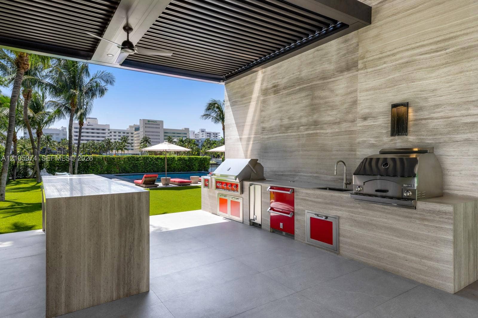 4395 Pine Tree Drive, Miami Beach, FL 33140 Covered outdoor kitchen featuring stone-clad surfaces, stainless steel built-in grill and beverage refrigeration, integrated faucet, and an overhead louvered pergola with a ceiling fan