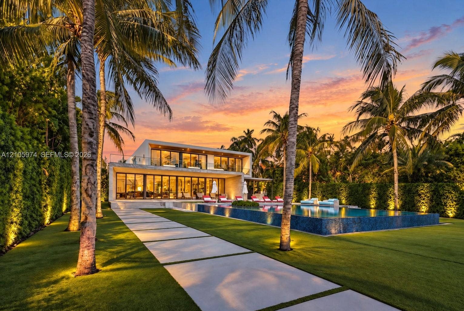 4395 Pine Tree Drive, Miami Beach, FL 33140 Modern architectural exterior featuring floor-to-ceiling glass wall systems, a second-story balcony with metal railings, a tiled infinity-edge pool, and concrete paver hardscaping
