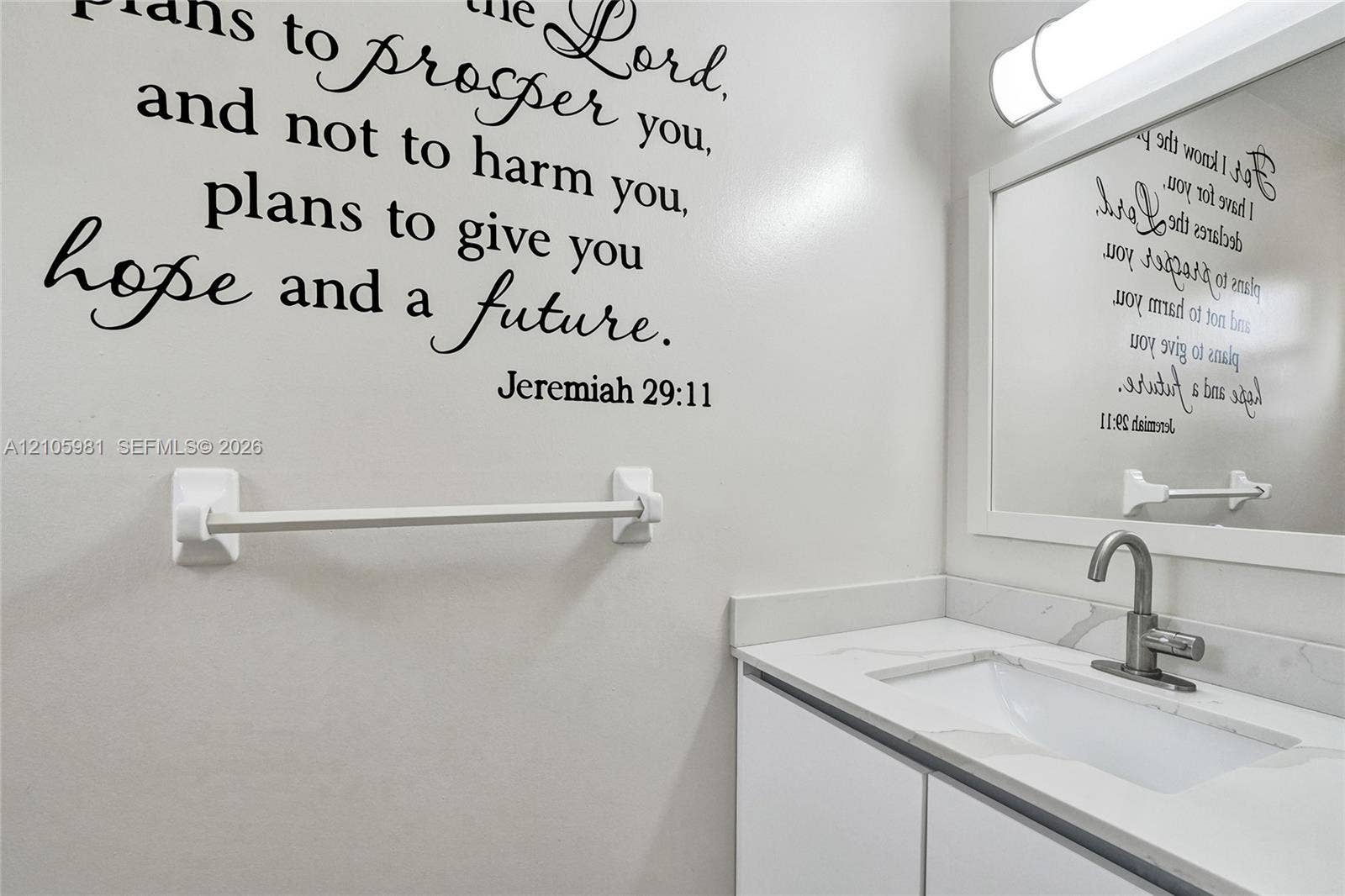 6813 NW 29th Avenue, Fort Lauderdale, Fort Lauderdale, FL 33309 Vanity featuring a white cabinet, light-toned countertop, integrated rectangular sink, and metallic faucet, accompanied by a wall-mounted light fixture, framed mirror, and white towel bar