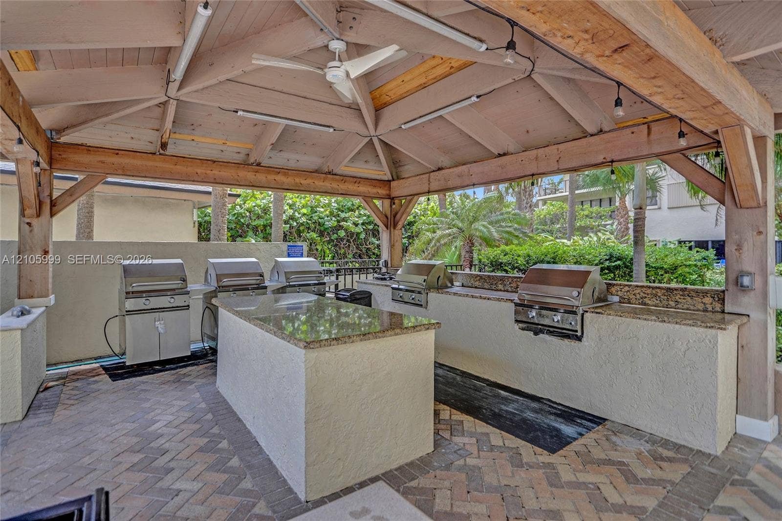 Covered outdoor kitchen featuring a timber-framed vaulted ceiling, stone countertops, multiple built-in stainless steel grills, and herringbone brick paver flooring