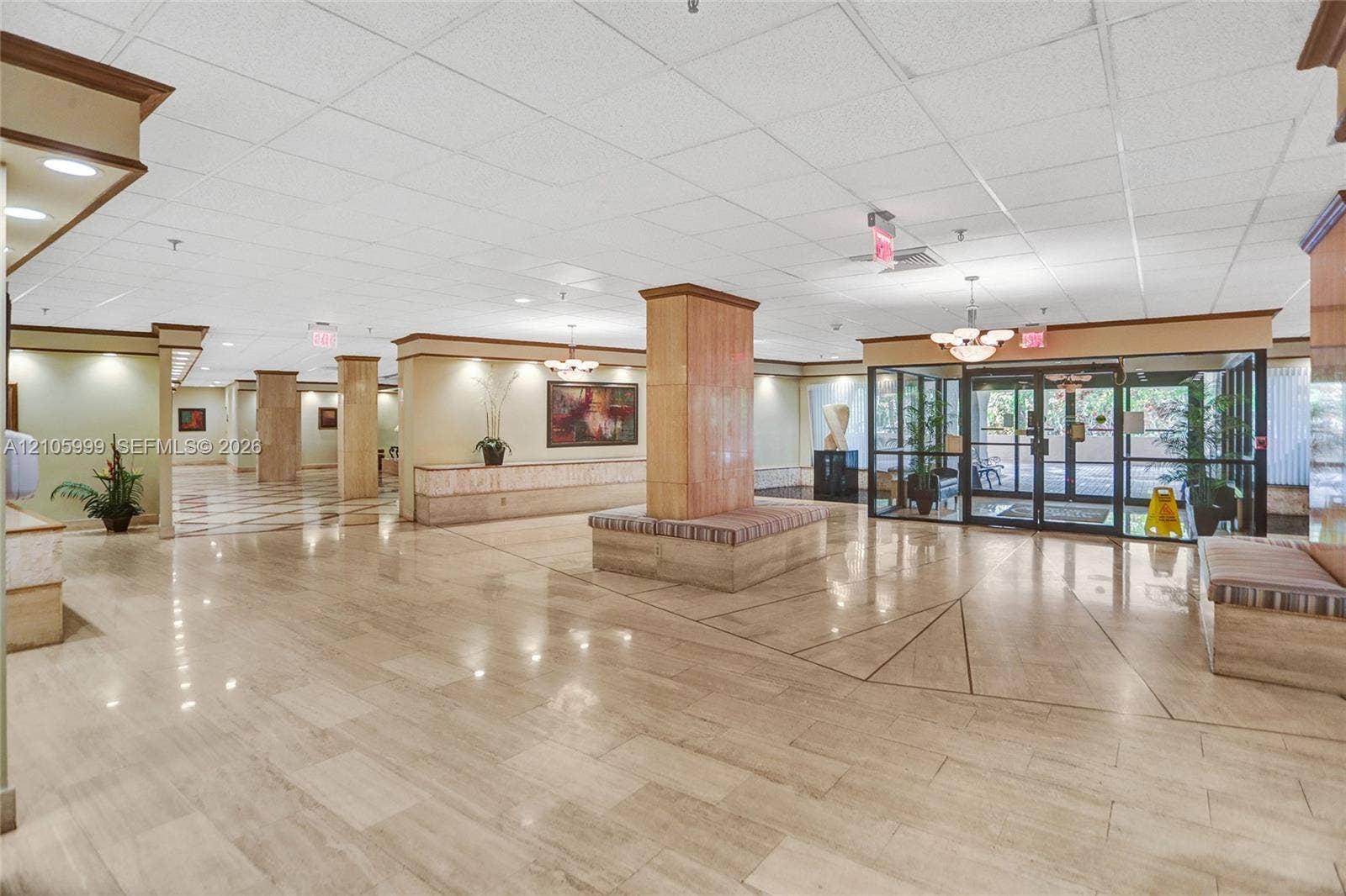 Spacious lobby featuring polished stone flooring, custom architectural columns with integrated seating, suspended acoustic tile ceiling, and glass entry doors